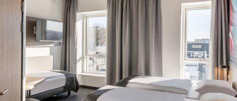 A hotel room with two beds, a desk, a flat-screen TV, and large windows with curtains.
