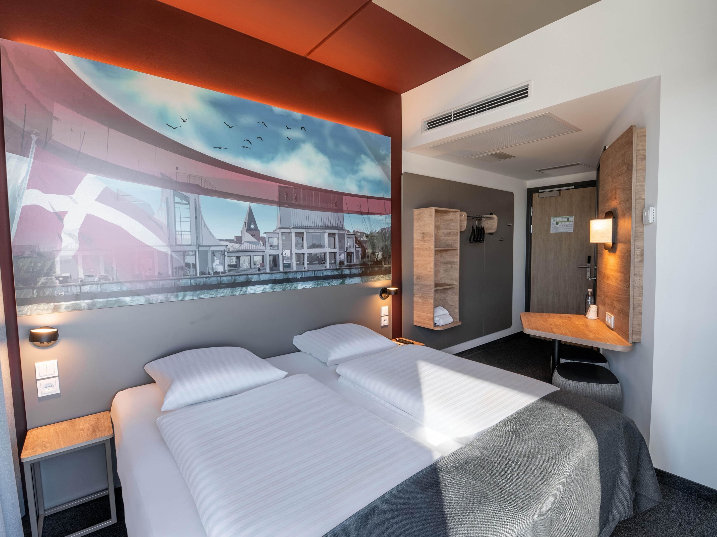 Habitación de hotel con una cama grande, cabecera con estante integrado, televisor montado en la pared, espejo y ventana con vistas a la ciudad.