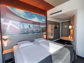 Habitación de hotel con una cama grande, cabecera con estante integrado, televisor montado en la pared, espejo y ventana con vistas a la ciudad.