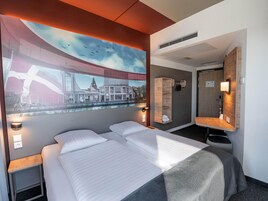 Habitación de hotel con una cama grande, cabecera con estante integrado, televisor montado en la pared, espejo y ventana con vistas a la ciudad.