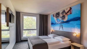 Une chambre d’hôtel moderne avec un grand lit, un téléviseur à écran plat, un tableau représentant des horloges et une vue sur les arbres par la fenêtre.