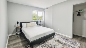 A bedroom with a bed, a nightstand, a lamp, a window, and a closet.