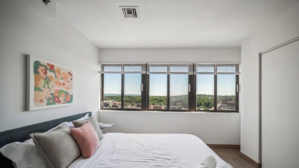 A modern hotel room with a large bed, a city view through floor-to-ceiling windows, a ceiling vent, and a piece of abstract art on the wall.