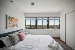 Een moderne hotelkamer met een groot bed, stadszicht door ramen van vloer tot plafond, een ventilatie rooster in het plafond, en een abstract kunstwerk aan de muur.