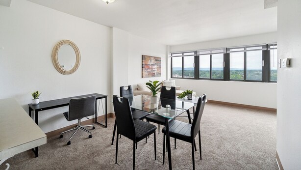 A modern office space with a glass table, black chairs, a white desk, a potted plant, and a large window offering a city view.