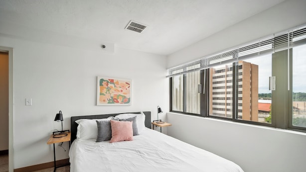 A modern hotel room with a large bed, a small bedside table with a lamp, a painting on the wall, and a window with blinds.