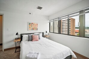 A modern hotel room with a large bed, a small bedside table with a lamp, a painting on the wall, and a window with blinds. - PP 501 Harrisburg Gem 2br, Pool, Parking (Harrisburg)