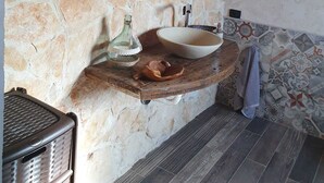Un mur en pierre surmonté d’un miroir rectangulaire et d’une étagère en bois où sont posés un lavabo et une bouteille.