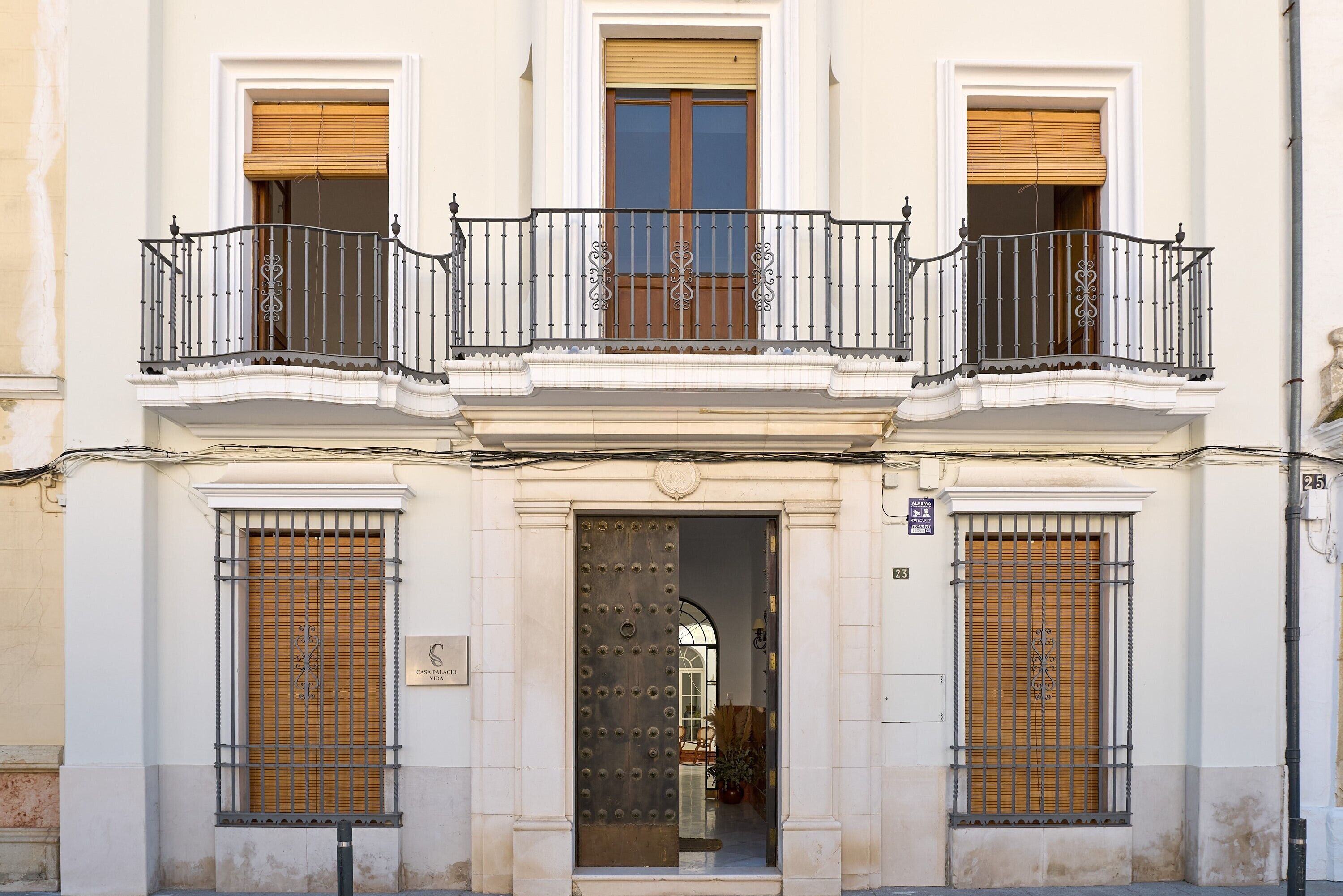 Un edificio con entrada central, dos balcones con barandillas de hierro forjado y una puerta de madera grande con barrotes.