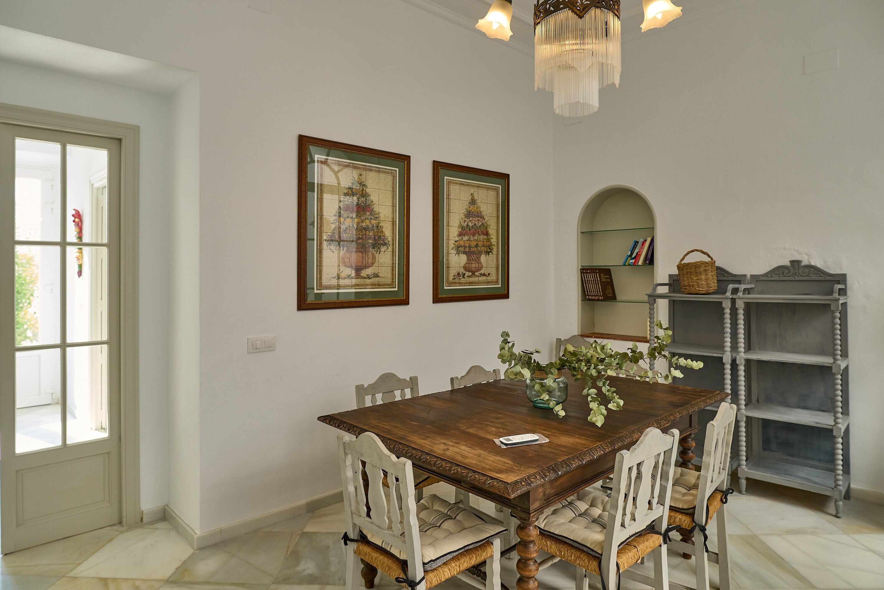 Comedor con mesa de madera, seis sillas, dos árboles de Navidad enmarcados, una araña y una estantería.