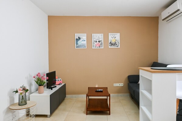 A modern living room with a beige wall, a white sofa, a small wooden table, a TV on a white cabinet, and three framed pictures.