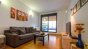 A modern living room with a sofa, coffee table, and a desk with a computer. There are three paintings on the wall, a large window, and a view of the outside. - Modern Family Apartment with Terrace and Pool in Madrid Tempory (Madrid)