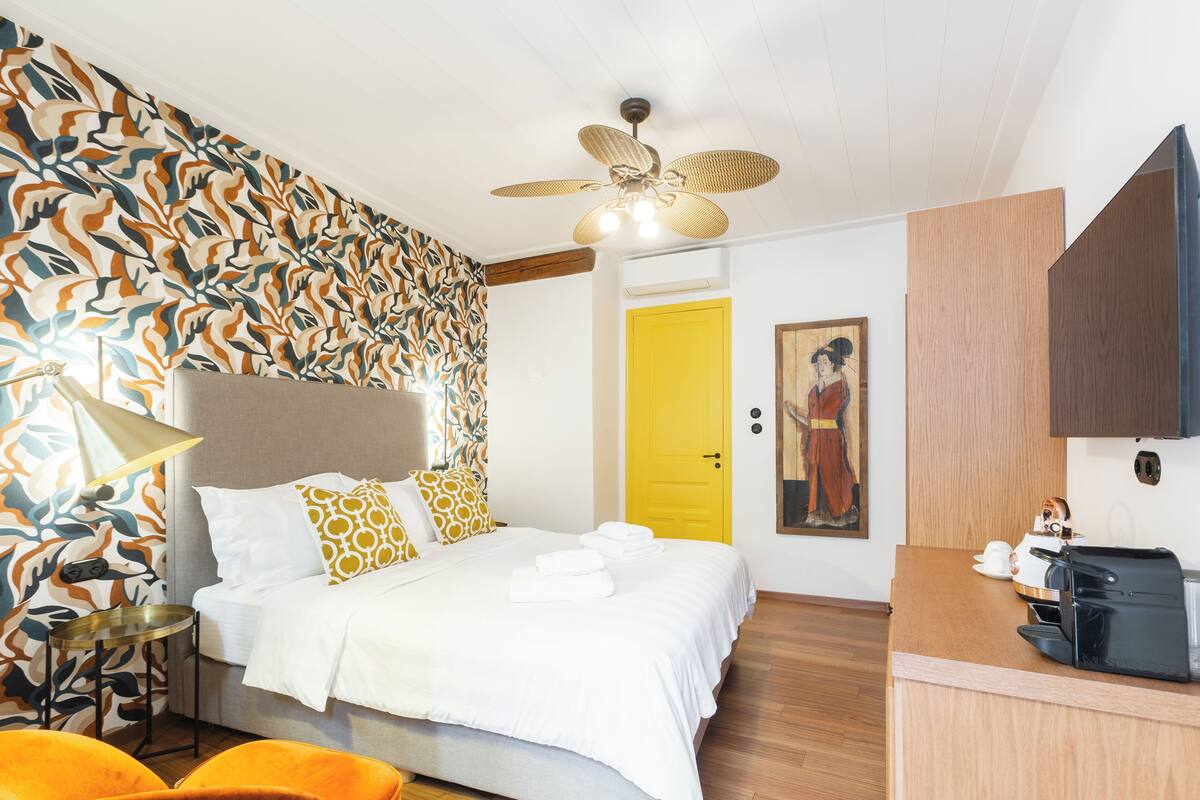 A hotel room with a bed, a desk, a chair, a ceiling fan, and a yellow door.