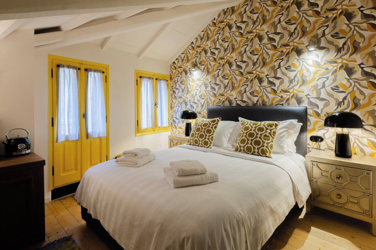 A hotel room with a bed, two bedside tables, a nightstand, a yellow door, and a patterned wall.