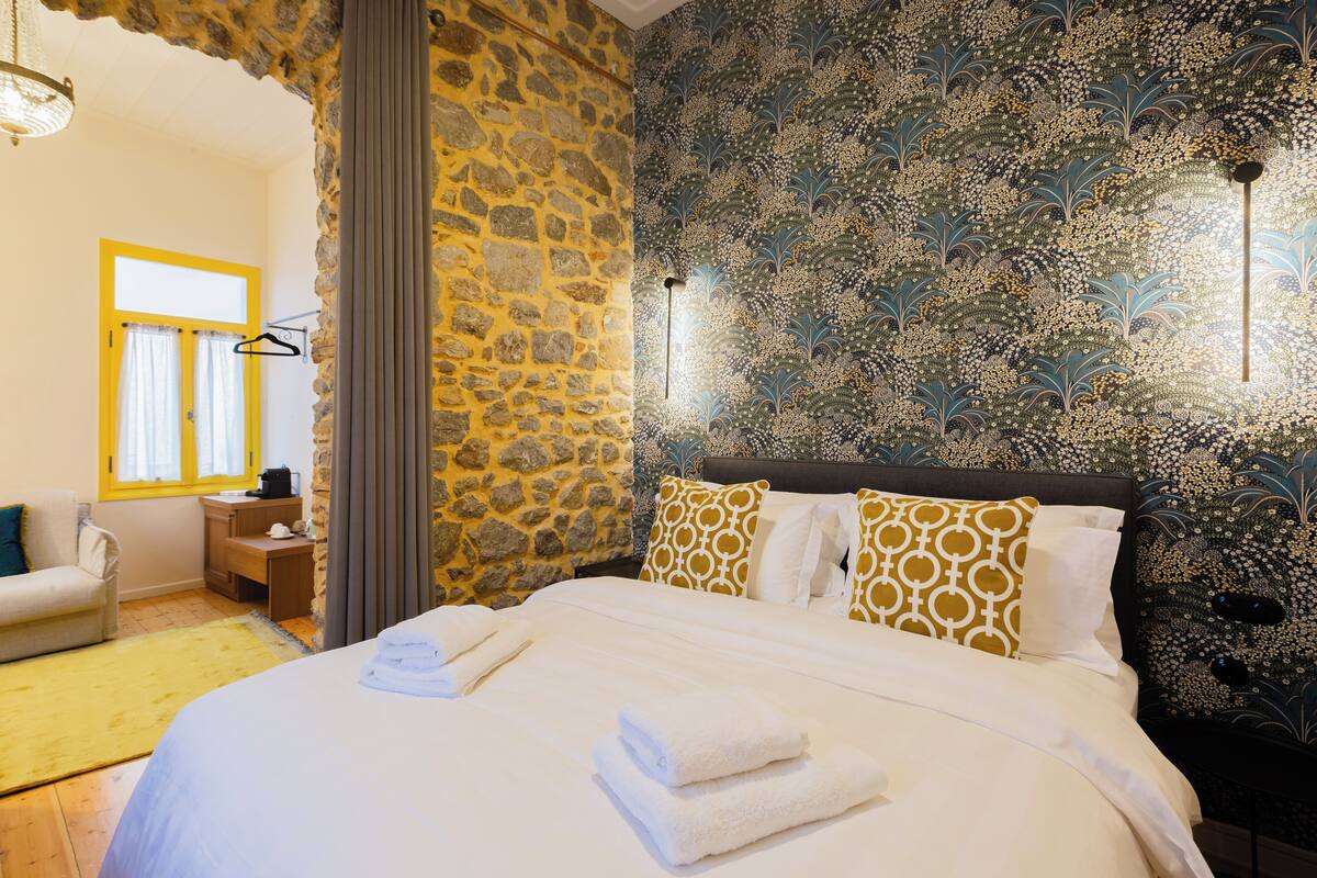 A bedroom with a stone wall, patterned wallpaper, a bed with white linens, a wooden nightstand, and a desk with a chair.