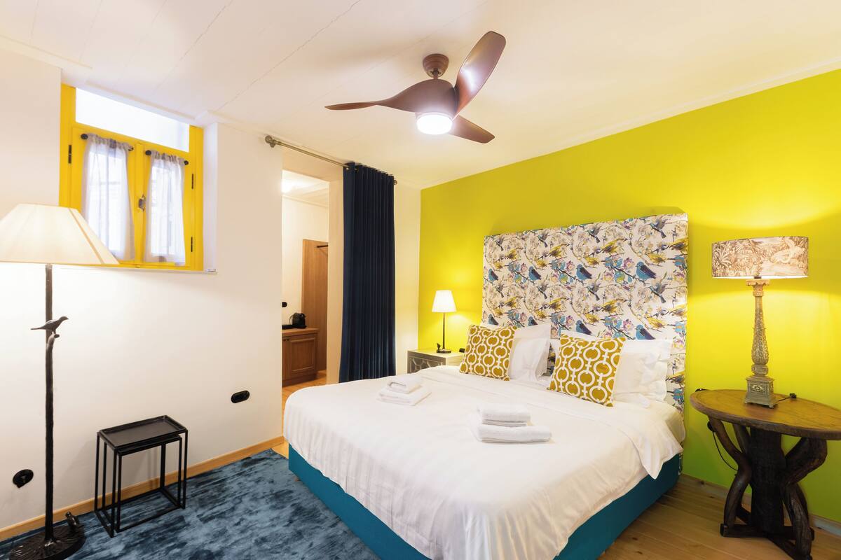 A hotel room with a bed, a ceiling fan, a nightstand, a lamp, and a window with curtains.