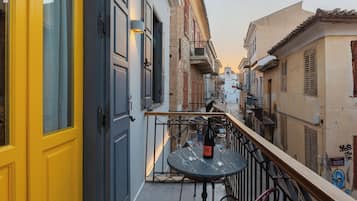 A narrow street with buildings on either side, a balcony with a table and wine bottle, and a yellow door.