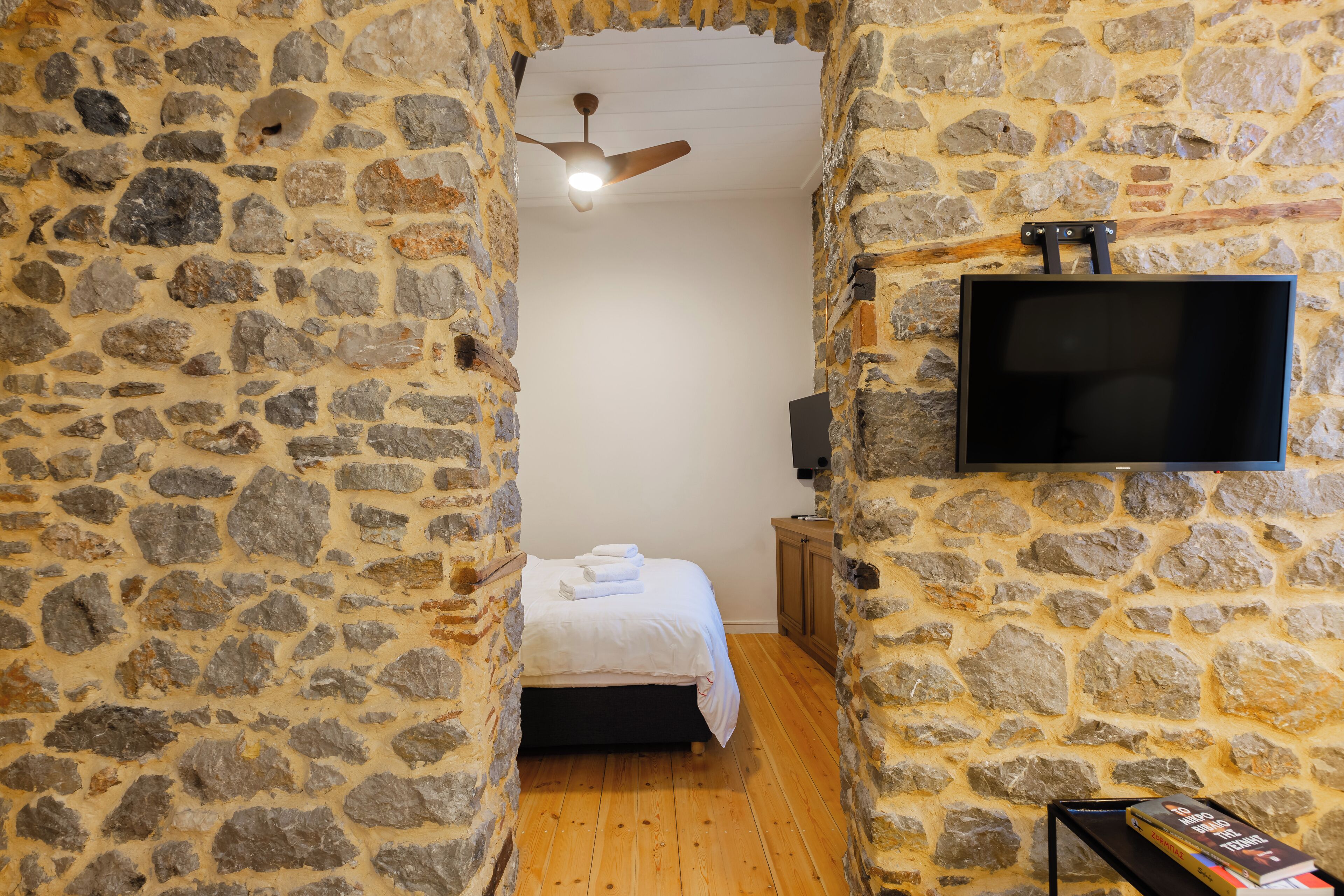 Une pièce aux murs de pierre, avec un lit, un ventilateur de plafond et un téléviseur fixé au mur.
