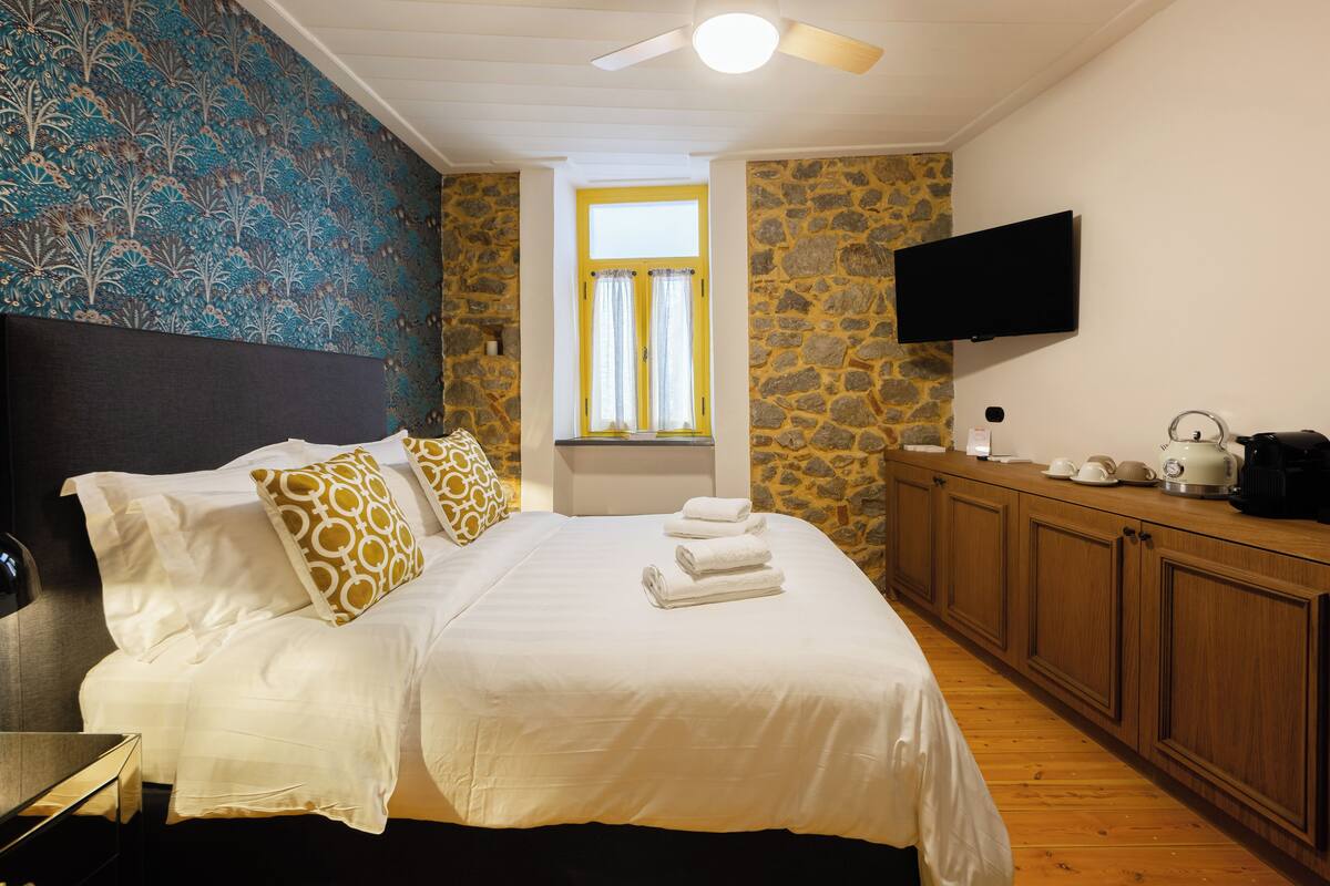 A hotel room with a bed, a flat-screen TV, a window with curtains, a stone wall, and wooden cabinets.