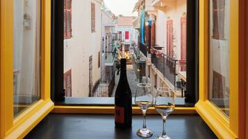 Une bouteille de vin et deux verres sont posés sur le rebord d’une fenêtre, d’où l’on aperçoit une rue étroite bordée d’immeubles.