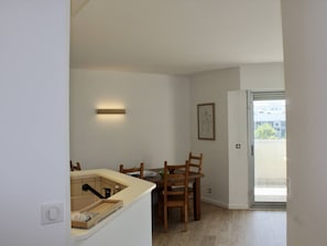 A compact kitchen with a built-in sink, wooden chairs, and a small dining table. A framed artwork is on the wall, and a sliding glass door leads to a balcony.