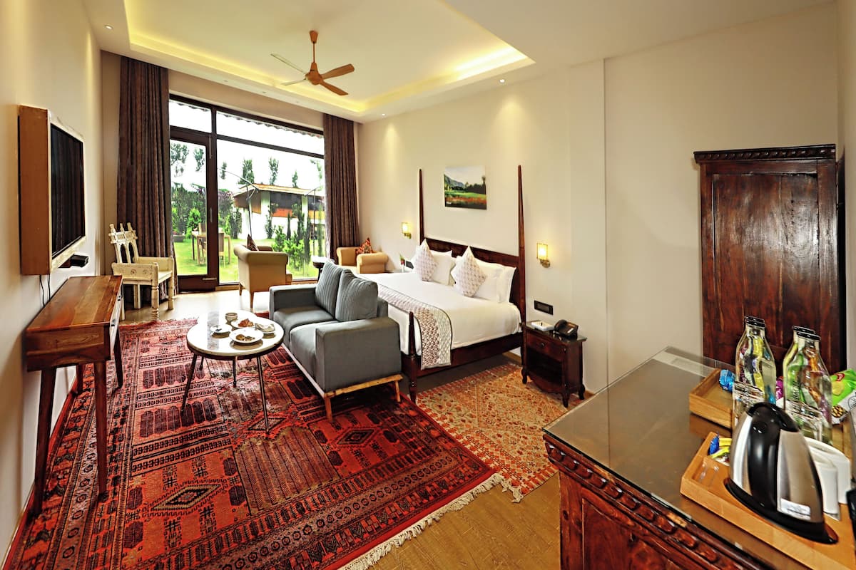 A hotel room with a bed, a sofa, a coffee table, a TV, a ceiling fan, and a large window with a view of greenery.