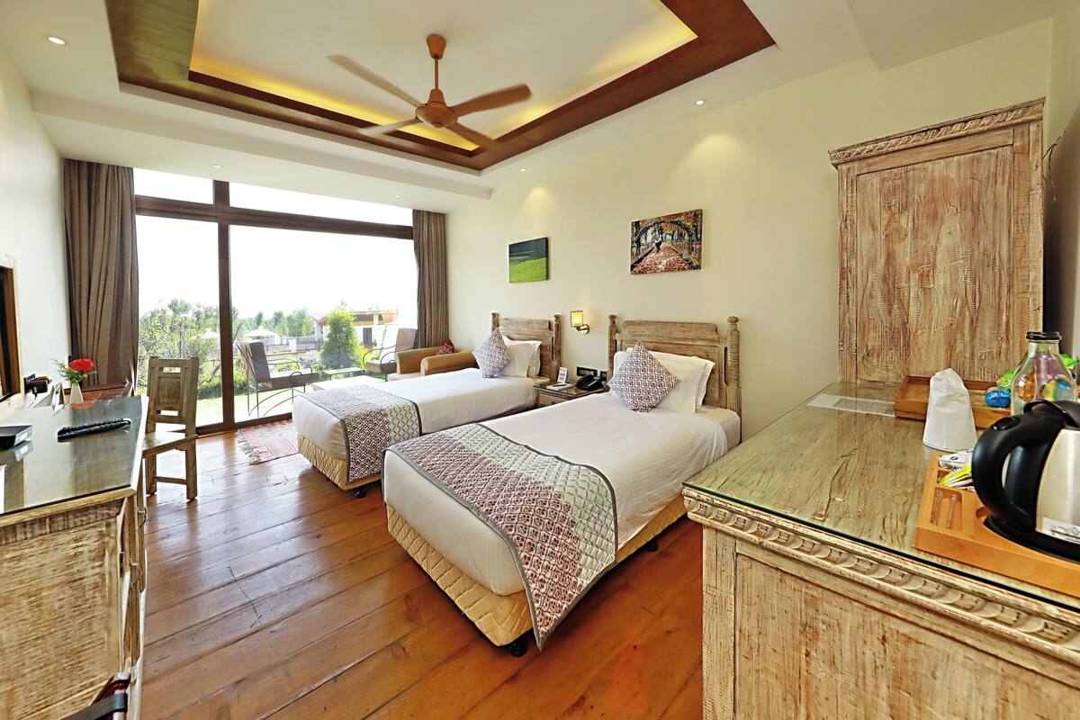 A hotel room with two beds, a dining table, a ceiling fan, and a large window with a view of the outdoors.