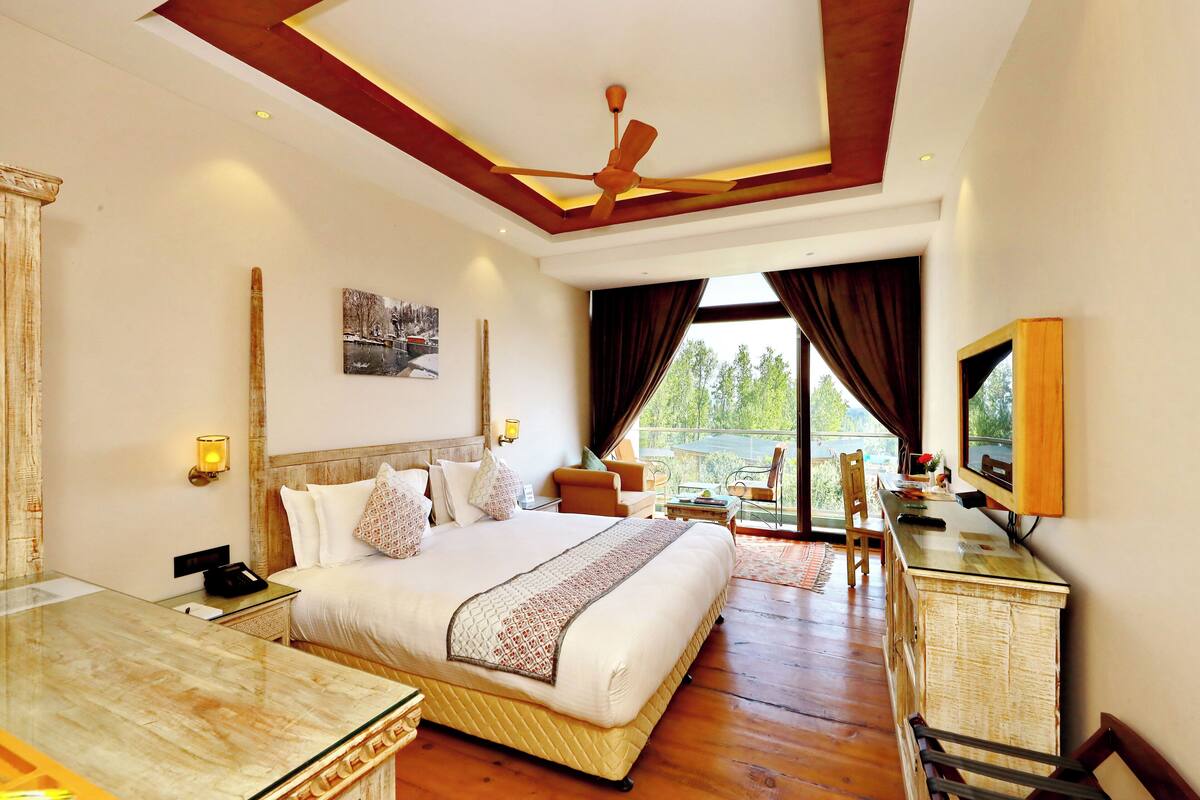 A hotel room with a large bed, a desk, a chair, a TV, and a balcony with a view.