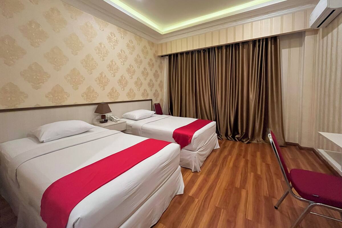 Habitación de hotel con dos camas, un escritorio, una silla, una mesita de noche con lámpara y una cortina.