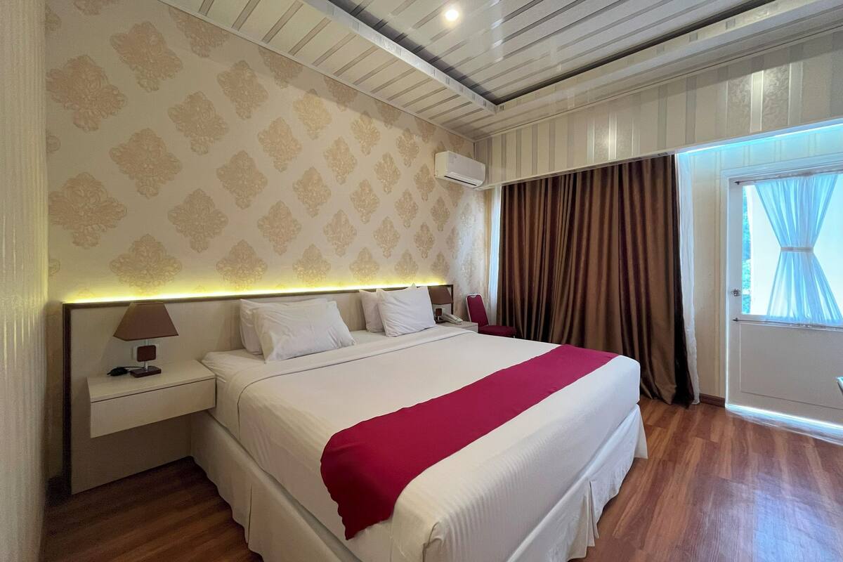 Una habitación de hotel con una cama grande, una mesita de noche con lámpara, aire acondicionado instalado en la pared y una ventana con cortinas.