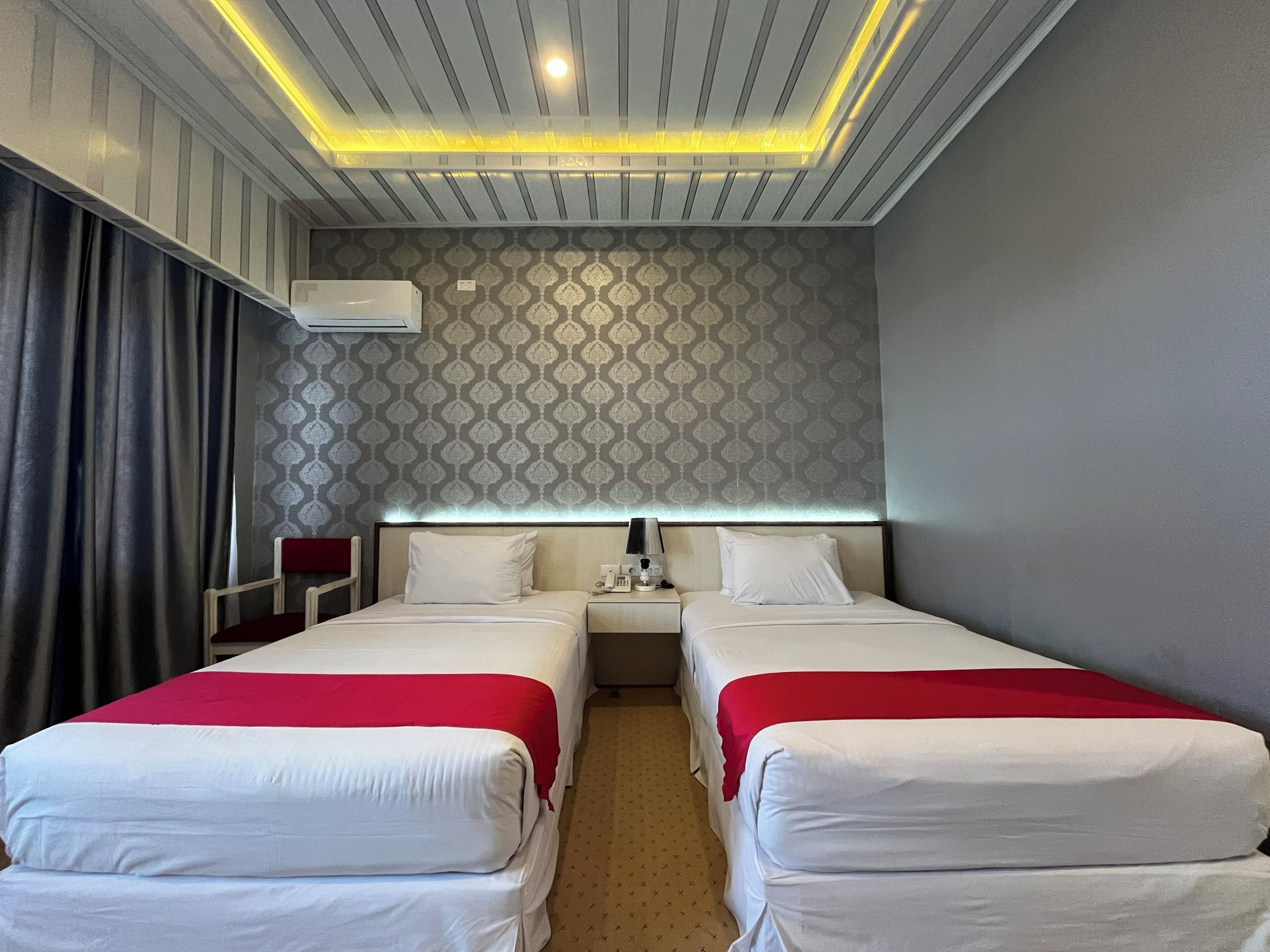 A hotel room with two beds, a bedside table with a lamp, an air conditioning unit, and a patterned wallpaper.
