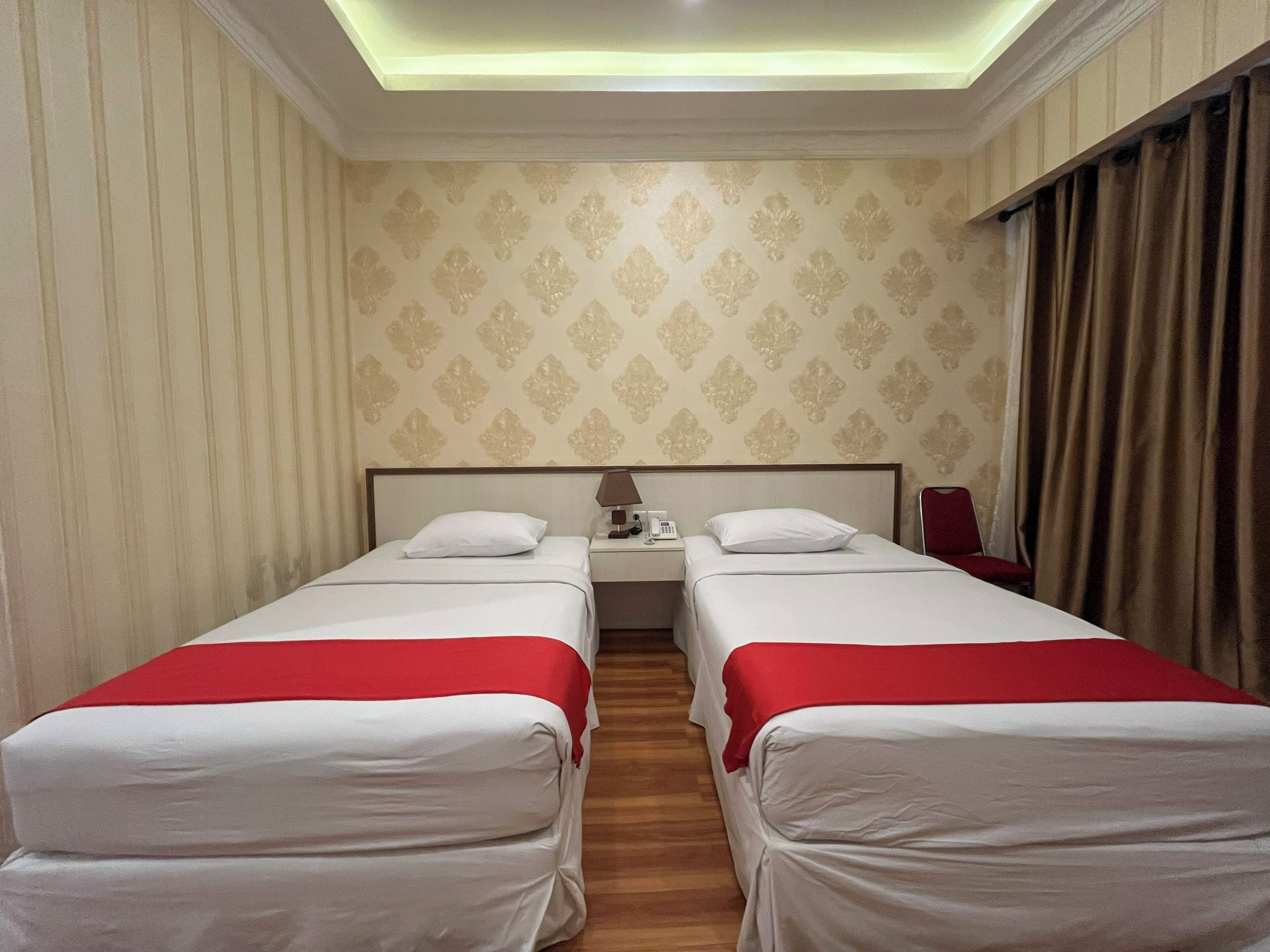 Une chambre d’hôtel avec deux lits, une table de chevet avec une lampe et un téléphone, et un fauteuil rouge.