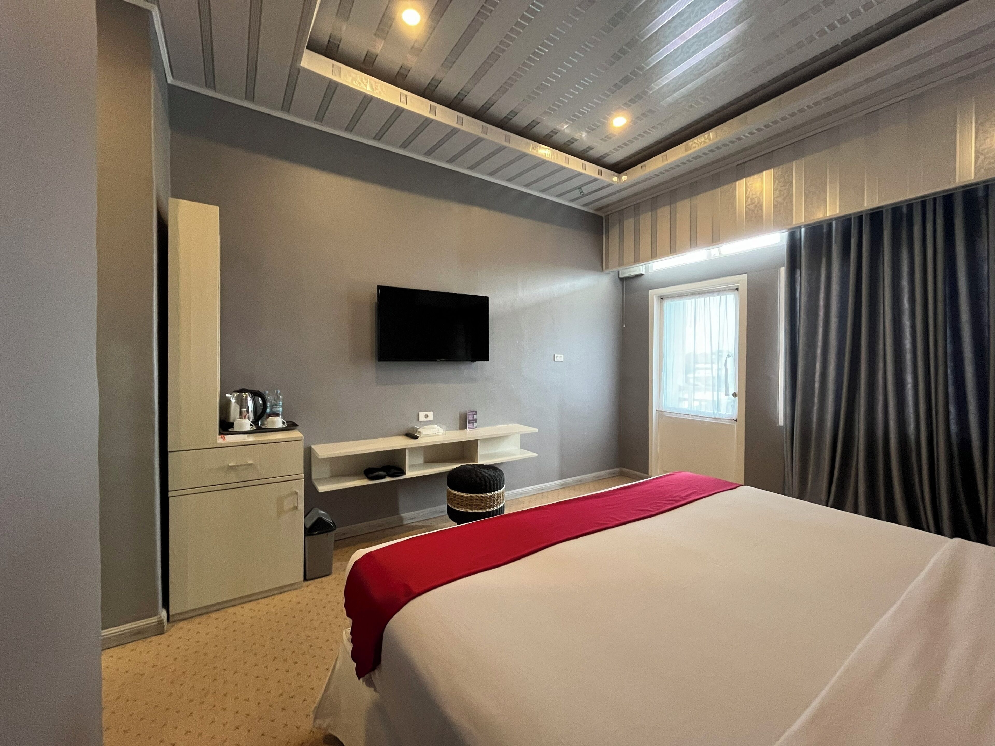 A hotel room with a bed, a TV mounted on the wall, a small desk with a chair, and a window with curtains.