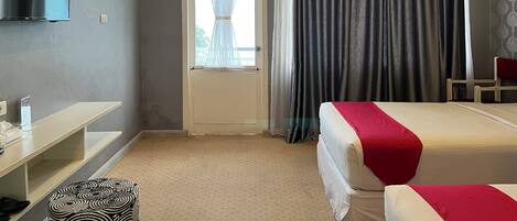 A hotel room with a bed, a TV, a desk, a chair, a window with curtains, and an air conditioning unit.