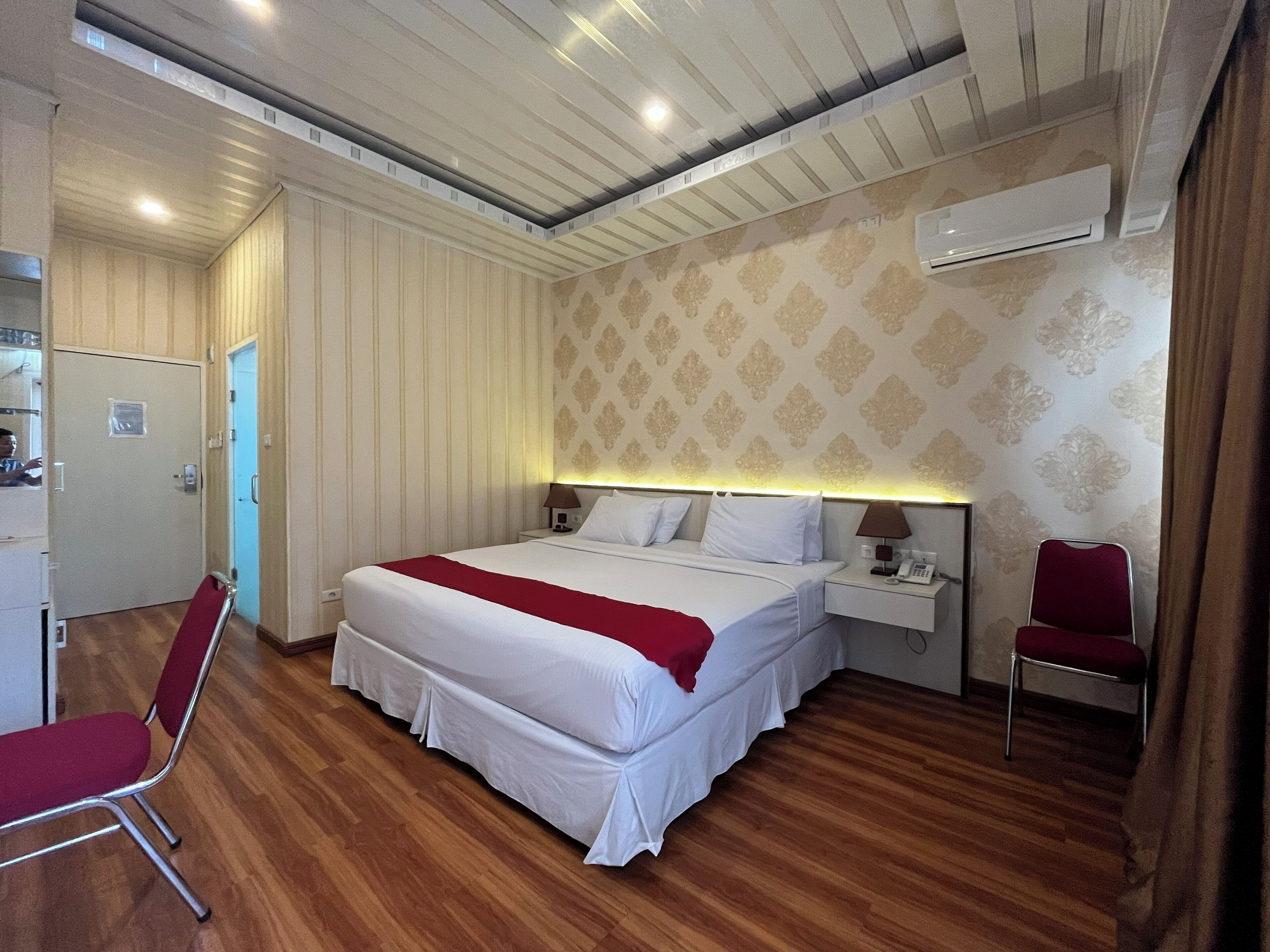 A hotel room with a bed, bedside tables, a chair, a wardrobe, and an air conditioning unit.