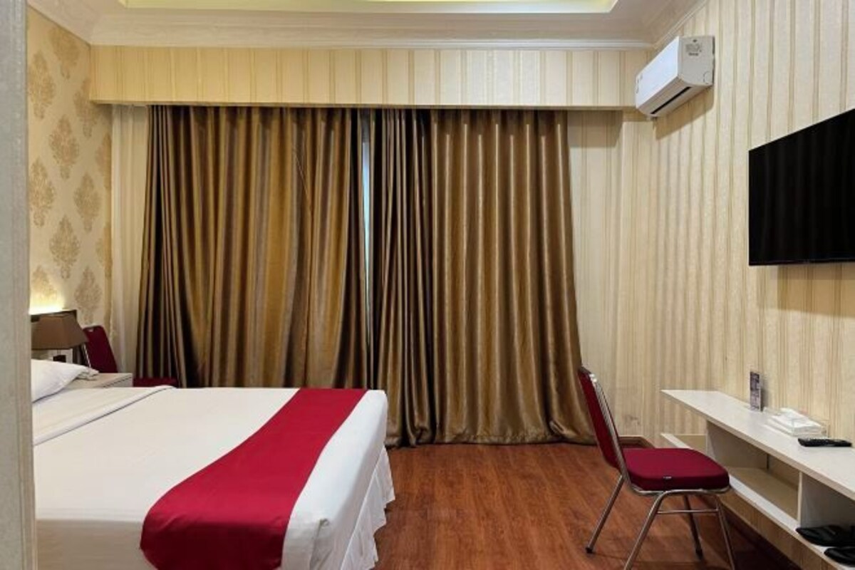 A hotel room with a bed, a desk, a chair, an air conditioning unit, and a television.