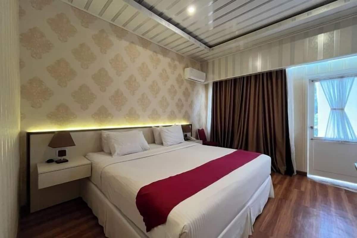 Una habitación de hotel con una cama grande, una mesita de noche, un equipo de aire acondicionado y una ventana con cortinas.