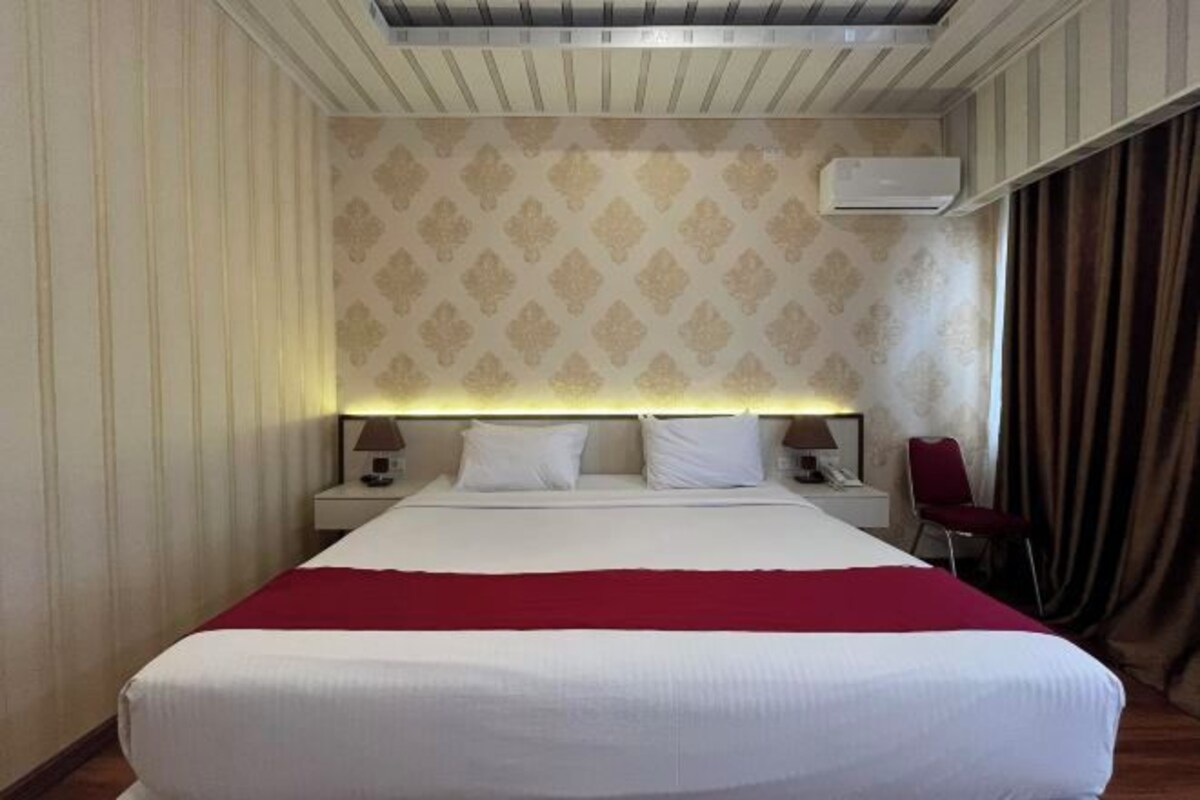 A hotel room with a large bed, a headboard with a light, two bedside tables with lamps, a red chair, and an air conditioning unit on the wall.