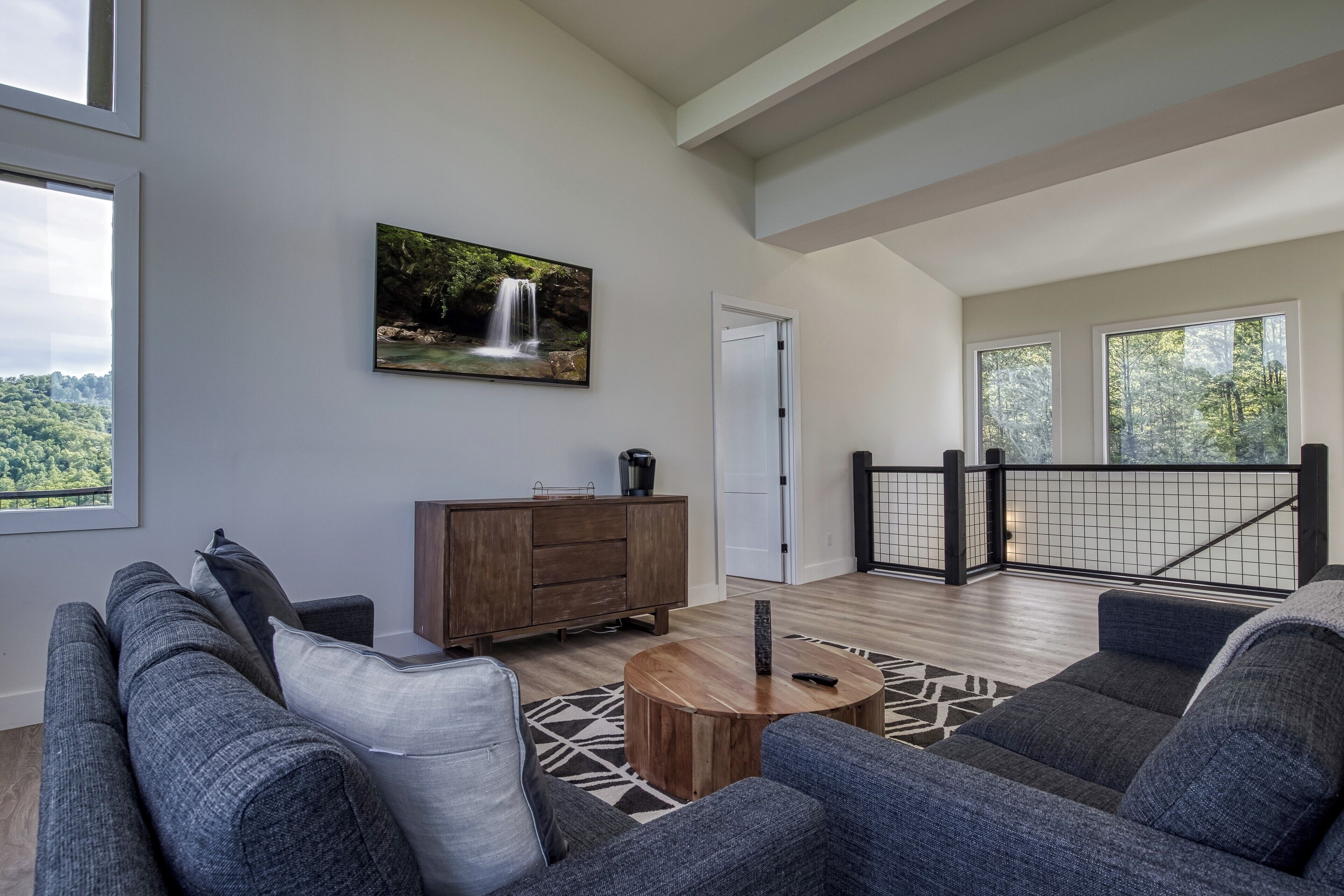 A spacious living room with a gray sofa, a wooden coffee table, and a painting of a waterfall.