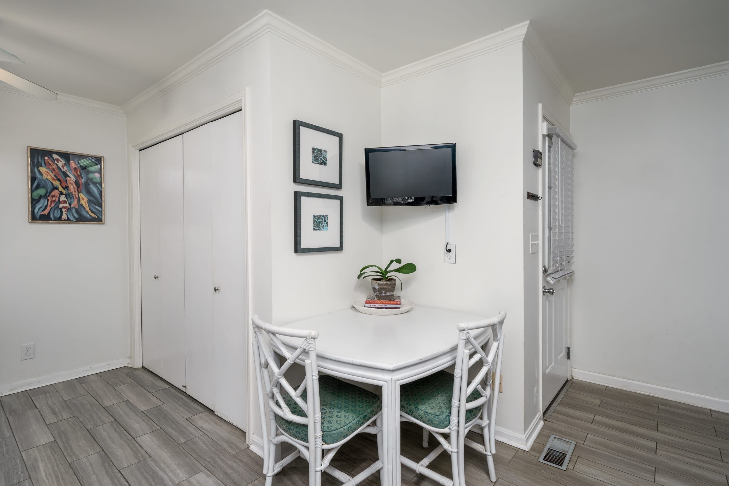 A corner room with a white wall, a flat-screen TV, two framed pictures, a small table with a plant, and a white door.