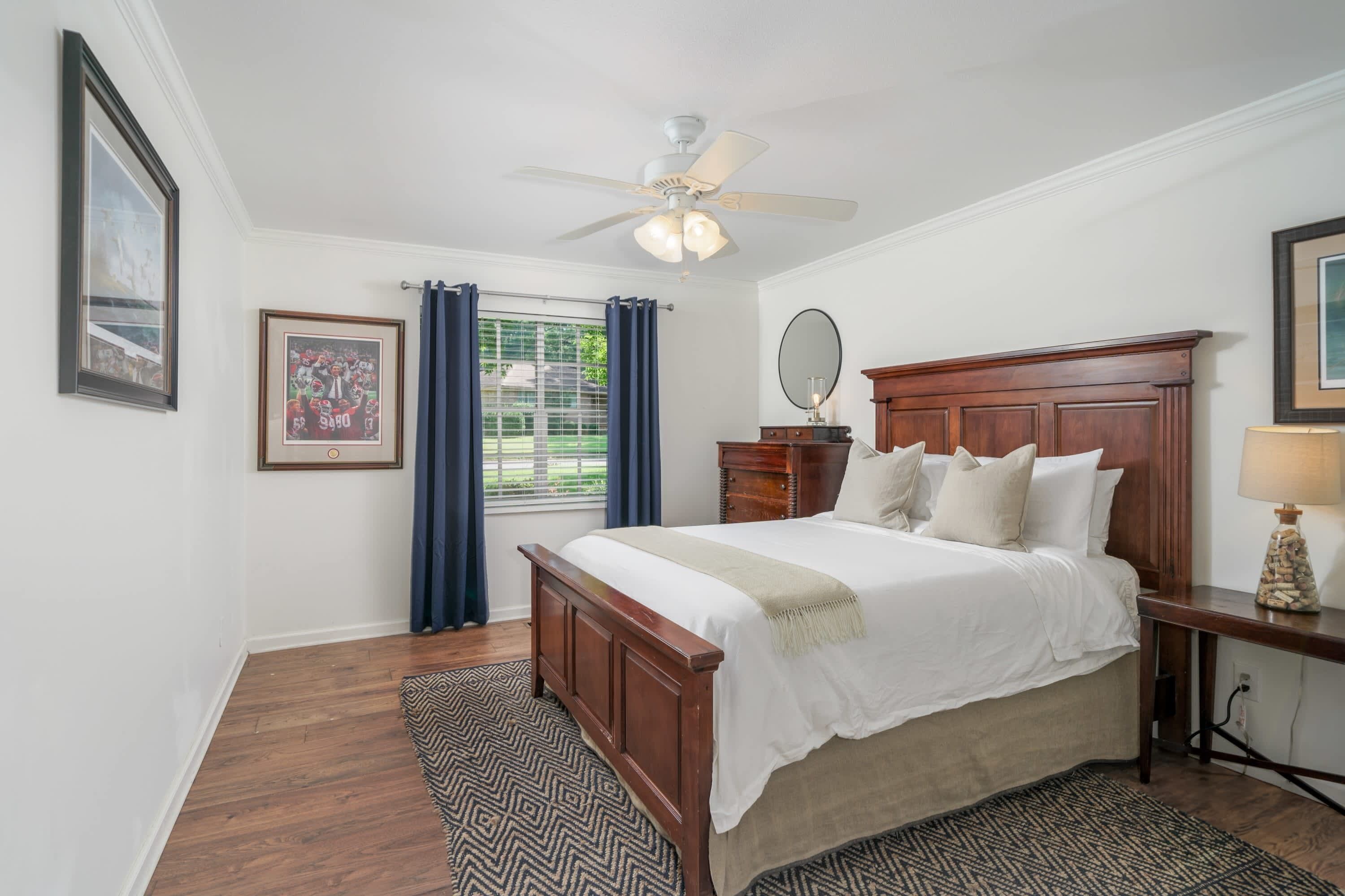 Une chambre avec un lit en bois, un ventilateur de plafond, une fenêtre avec des rideaux et un tableau encadré au mur.