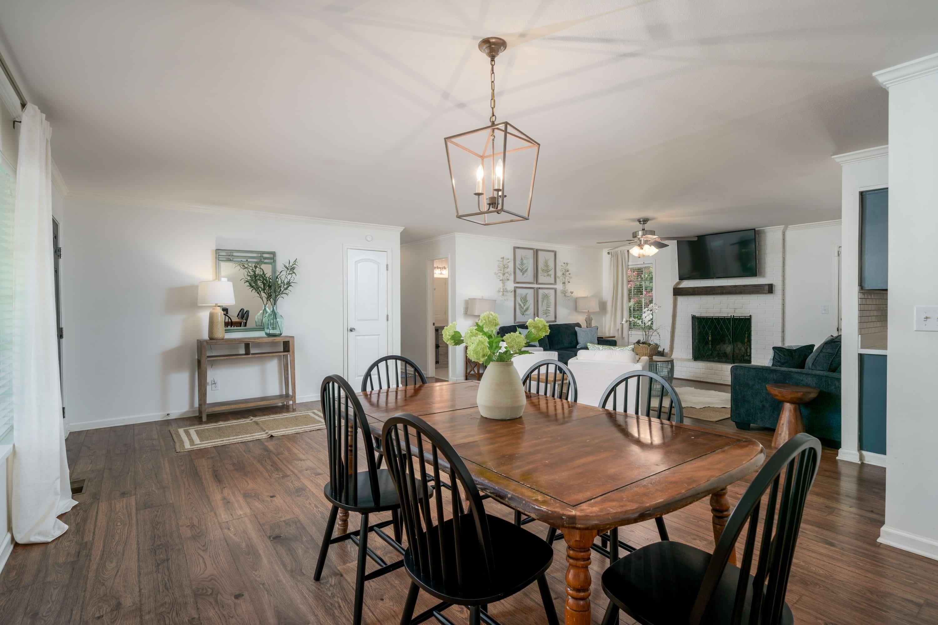 A spacious dining area with a wooden table and chairs, a hanging light fixture, and a fireplace in the background.