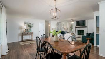 A spacious dining area with a wooden table and chairs, a hanging light fixture, and a fireplace in the background.