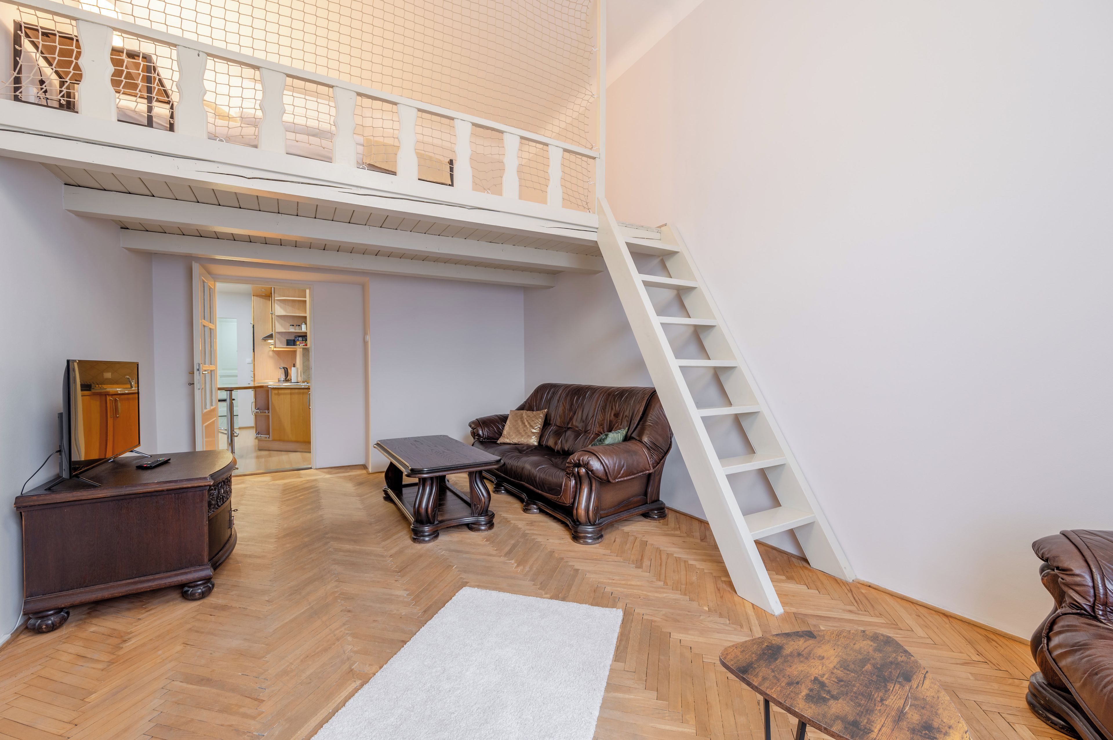 Een ruime loft met een hoge nok, een houten visgraatvloer, een leren bank, een salontafel, een televisie op een houten kast en een witte trap.