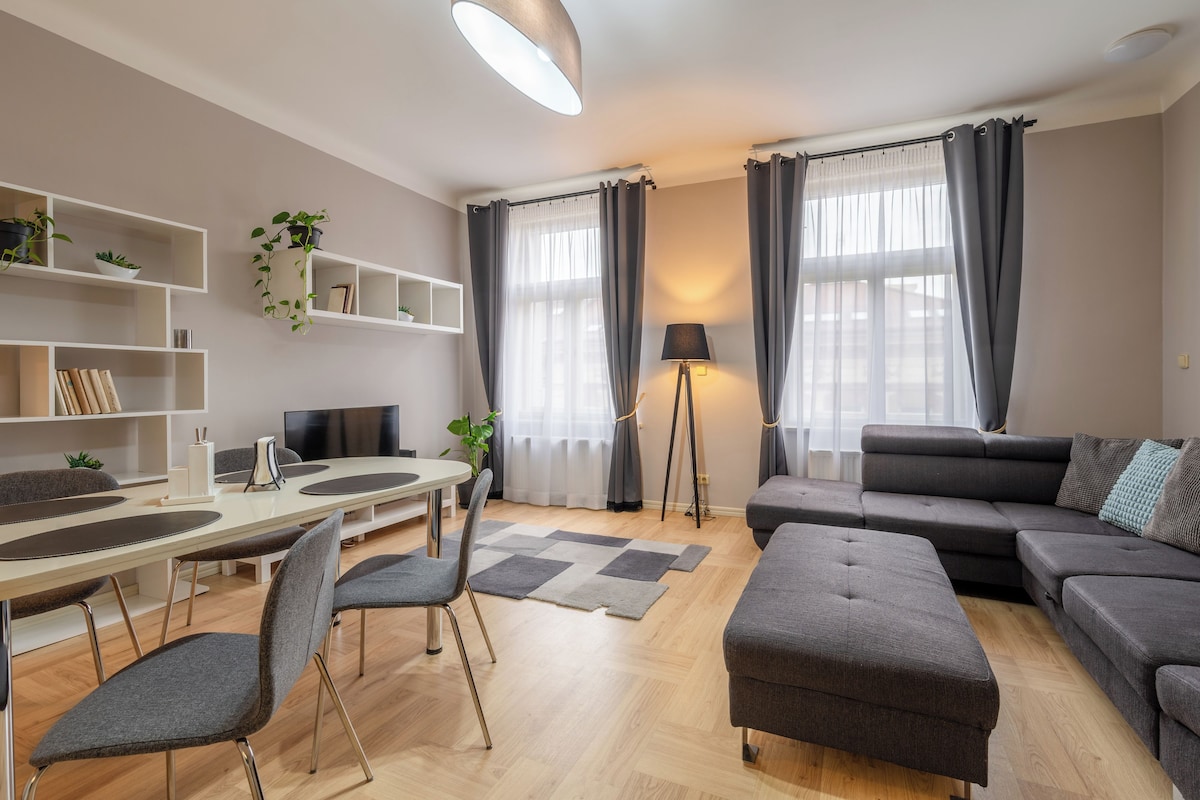A modern living room with a dining area, grey sofas, a coffee table, a TV, and a bookshelf with plants.