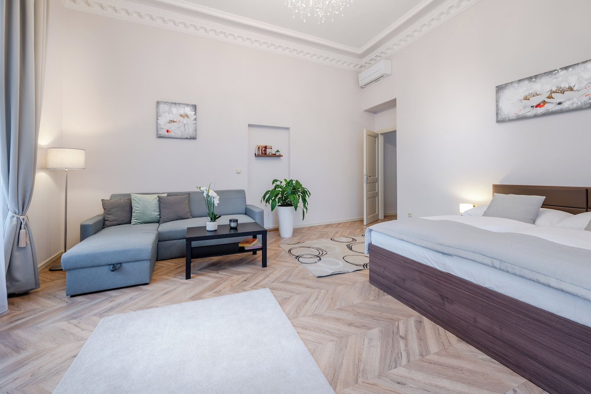 A modern hotel room with a grey sofa, a bed, a coffee table, and a potted plant.