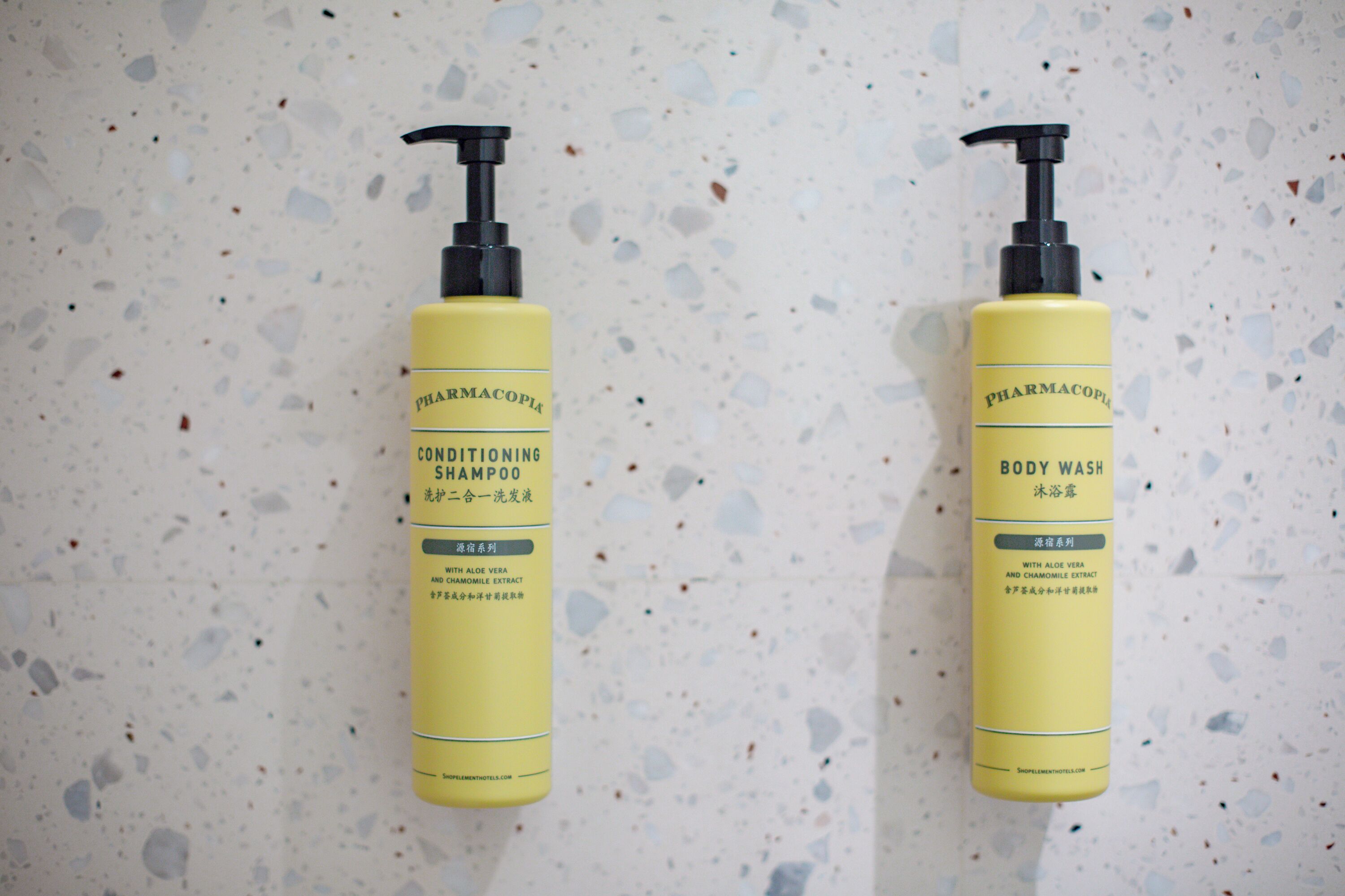 Two yellow bottles of Pharmacopia body wash and conditioning shampoo on a speckled surface.