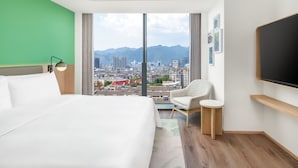 Une chambre d’hôtel avec un grand lit, une chaise, un téléviseur et une vue sur un paysage urbain, avec des montagnes en arrière-plan.