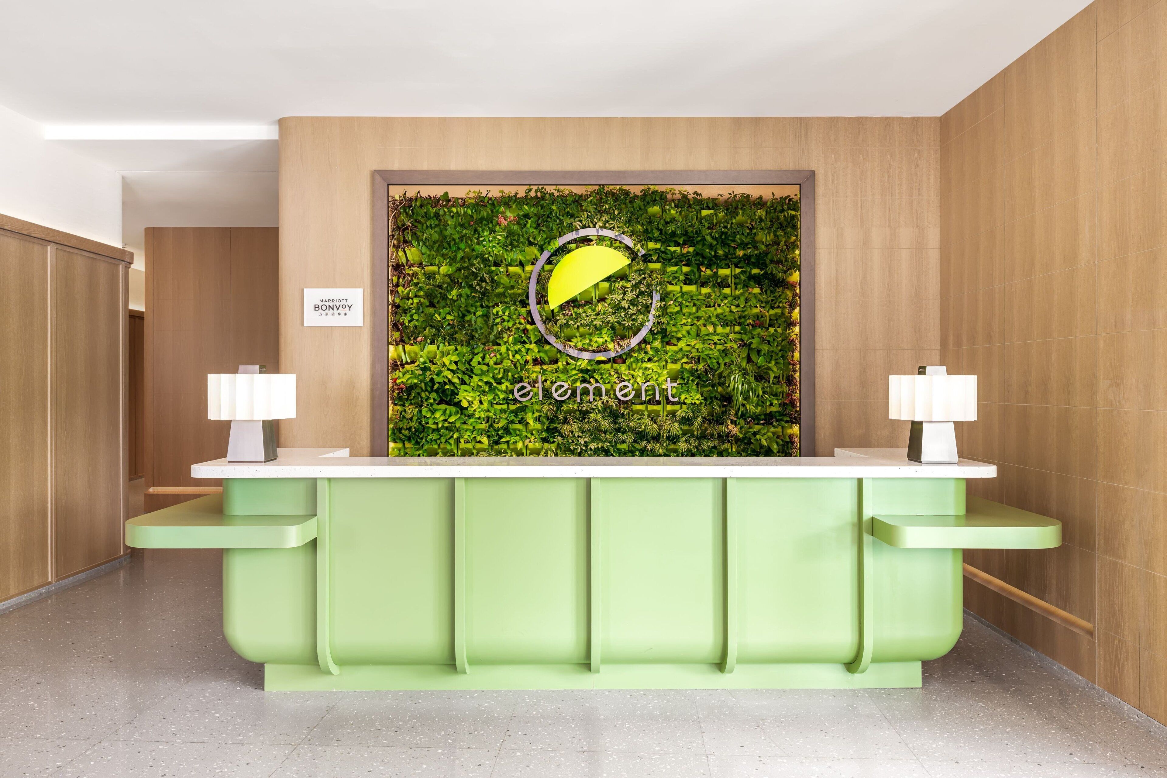 Un mostrador de recepción con una encimera verde y dos lámparas, una pared con un artístico revestimiento de hojas verdes, y un cartel que dice “element”.