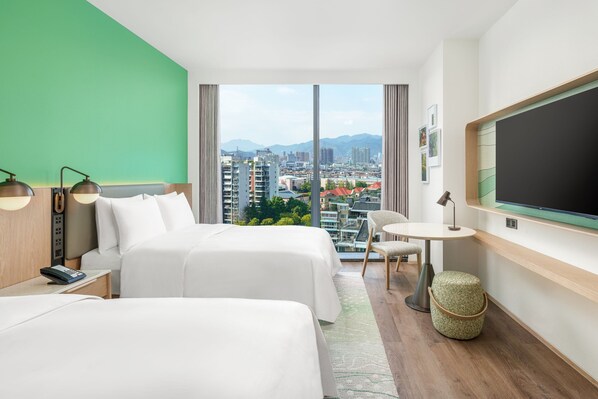 A hotel room with two beds, a small table, a chair, a TV, and a view of the city and mountains. - Element Lishui (Lishui)